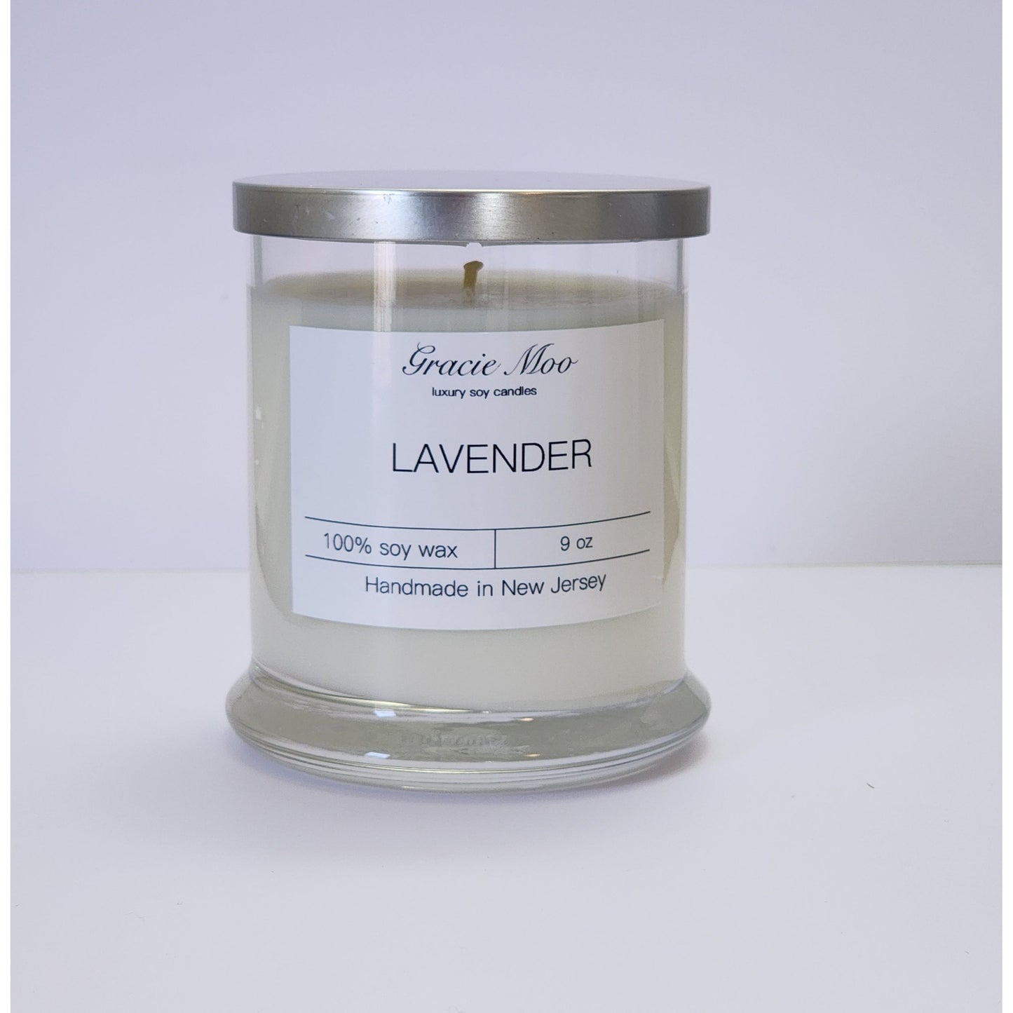 Lavender Status Candle by Gracie Moo Candles with a tall white candle in a glass holder, surrounded by lavender flowers.